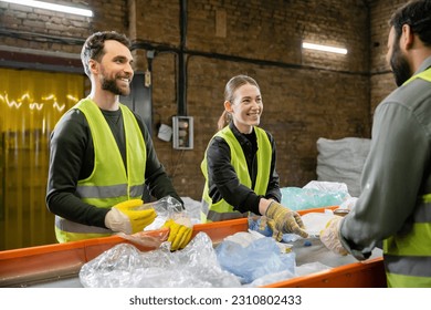 Cheerful sorters in vests and protective gloves sorting garbage and looking at blurred indian colleague near conveyor in waste disposal station, garbage sorting and recycling concept - Powered by Shutterstock