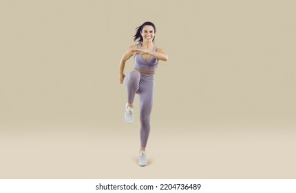 Cheerful sportswoman performs sport jumps raising her knees high on beige background. Full length portrait of smiling energetic woman in sportswear showing sports exercises. Banner. Isolated.