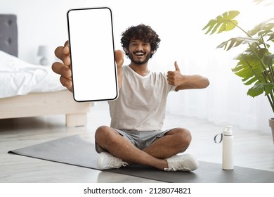 Cheerful Sporty Millennial Indian Guy With Beard And Curly Hair Sitting On Yoga Mat, Showing Modern Smartphone With Empty Screen, Recommending Nice Fitness Mobile App, Bedroom Interior, Mockup