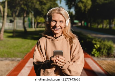 Cheerful Sporty Slim Mature Woman Female Runner Athlete Choosing Sound Track On Smart Phone While Listening To The Music Radio Podcast While Training Jogging In Stadium