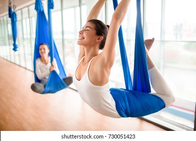 Cheerful sporty woman wearing top and leggings performing anti-gravity yoga exercise at spacious health club with panoramic windows