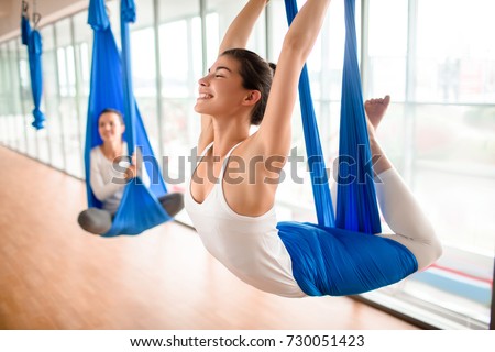 Cheerful sporty woman wearing top and leggings performing anti-gravity yoga exercise at spacious health club with panoramic windows