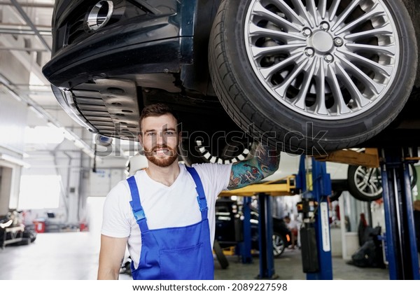 Cheerful strong happy repairman young male\
professional technician mechanic man wears blue overalls white\
t-shirt stand near car lift changes wheel tires work in vehicle\
repair shop workshop\
indoor
