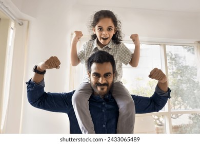 Cheerful strong Indian dad lifting little daughter kid on shoulders, making power hands gestures, showing fists, muscles, looking at camera with martial scream, playing active games - Powered by Shutterstock