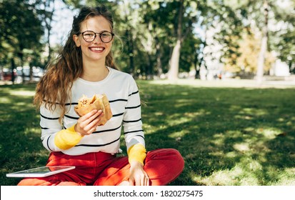 Cheerful Student Female In Casual Outfit, Glasses, Sitting On The Green Grass At The College Campus, Have Lunch, And Studying Outdoors. Smiling Woman Takes A Rest Eating Fast Food And Learning Outside