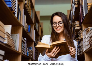 Cheerful student-girl, standing between bookshelves and reading a book. Express positive emotions while reading, intellectual leisure time.