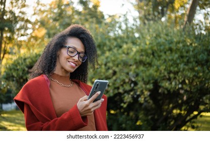 Cheerful Stylish Afro Hairstyle Black Woman Looking At Her Smartphone Outside In Autumn.