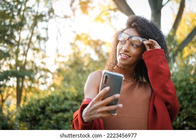 Cheerful Stylish Afro Hairstyle Black Woman Looking At Her Smartphone Outside In Autumn.