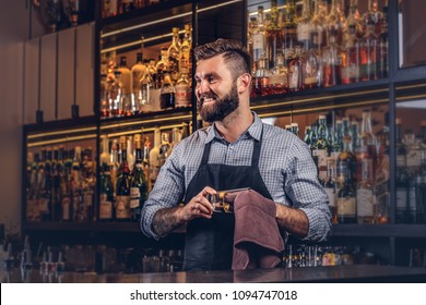 Cheerful stylish brutal barman is cleaning the glass with a cloth at bar counter background. - Powered by Shutterstock