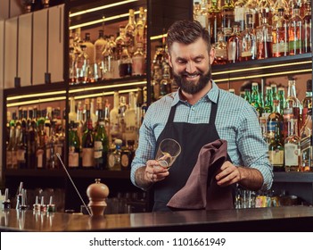 Cheerful stylish brutal barman is cleaning the glass with a cloth at bar counter background. - Powered by Shutterstock