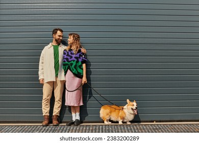 cheerful and stylish couple walking with corgi dog near grey garage door, animal companions - Powered by Shutterstock