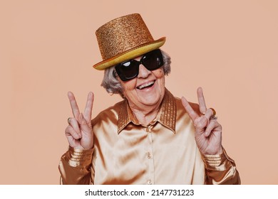 Cheerful stylish elderly grandmother wears gold hat, golden shirt and sunglasses, doing peace victory sign and celebrating party. Portrait at studio. Senior old woman looking at camera.