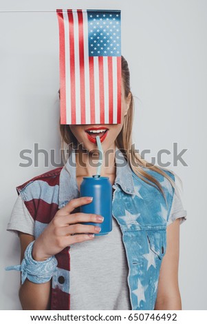 cheerful stylish girl in american patriotic outfit drinking soda from can, with american flag in front of her face isolated on grey 