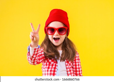 Cheerful Stylish Little Girl Dressed In Pink Checkered Shirt, Red Cap, Sunglasses, Jeans On Yellow Background. Smiling Cute Child Is Showing With Fingers Peace Sign, Number Two. Emotional Portrait.