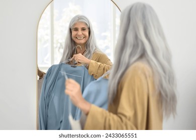 Cheerful stylish senior Latin woman choosing cloth for wearing at mirror, holding hanger with oversized jumper, looking at mirror, smiling, laughing, trying on new cloth - Powered by Shutterstock