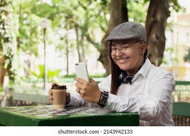 Cheerful Stylish Woman With Cup Of Coffee Looking At Cell Phone Screen Outside In Park And Woods. Technology, Social Media, Connection Concept.