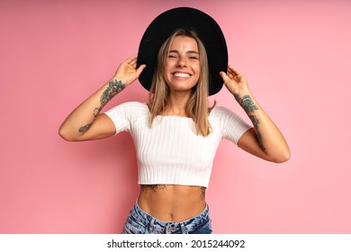 Cheerful stylish woman posing at studio pink background, wearing crop top blue jeans, black fedora. Blonde hairs and tattos. - Shutterstock ID 2015244092