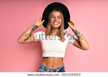 Cheerful stylish woman posing at studio pink background, wearing crop top blue jeans, black fedora. Blonde hairs and tattos.