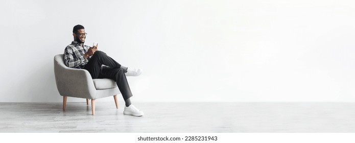 Cheerful successful african american millennial businessman sitting in chair and typing on smartphone on white wall background on empty space in office. Business with gadget for work, new normal - Shutterstock ID 2285231943