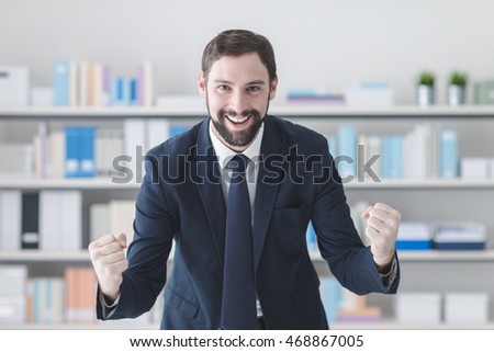 Cheerful successful businessman smiling in his office with raised fists, winning and enthusiasm concept