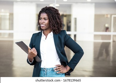 Cheerful successful manager with documents posing outside. Young black business woman standing at glass wall, holding folder, looking away, smiling. Successful professional concept