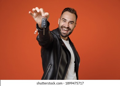 Cheerful successful young bearded man 20s wearing basic white t-shirt, black leather jacket standing hold in hands car keys looking camera isolated on bright orange colour background studio portrait