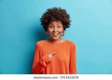 Cheerful surprised young pretty African American woman points at herself asks who me smiles broadly didnt expect being chosen dressed in long sleeved orange jumper isolated over blue background