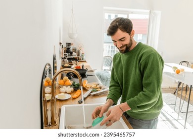 Cheerful and tattooed bearded man in green jumper and jeans washing plate at home - Shutterstock ID 2395465757