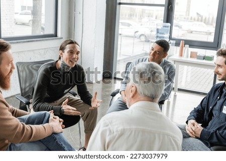 Cheerful tattooed man with alcohol addiction talking to interracial group during therapy in rehab center