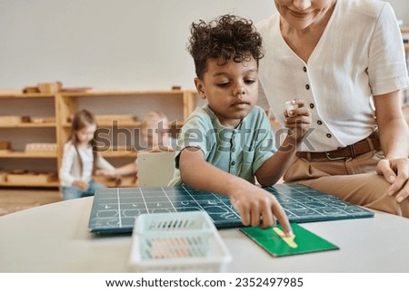 cheerful teacher observing african boy pointing at number near chalkboard, learning through play