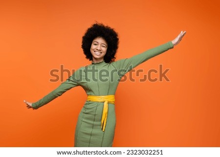 Cheerful and teen african american girl in stylish green dress and bold makeup winking and looking at camera while standing isolated on orange, teenage fashion and generation z concept