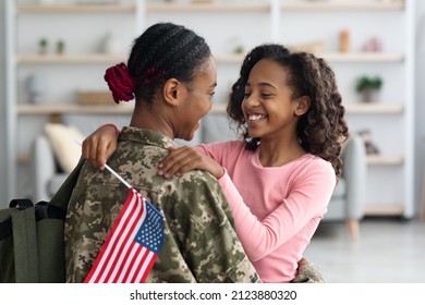 Cheerful Teen Black Girl With Flag Of The US Hugging Her Mom Wearing Camouflage Uniform, Daughter Looking At Mother Face And Smiling, Woman Coming Back Home From Military Service, Side View