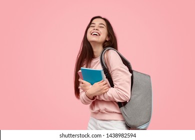 Cheerful teen girl with backpack embracing notepads and laughing while standing against pink background during studies