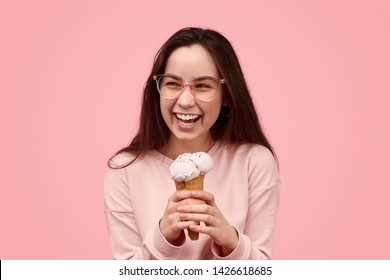 Cheerful teen girl in stylish glasses ice cream laughing and looking away while enjoying delicious ice cream against pink background