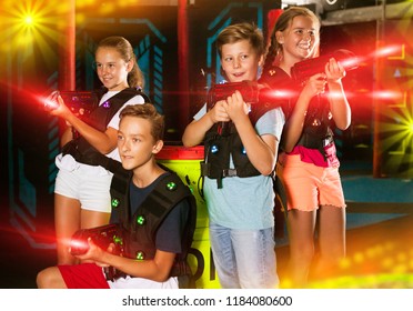Cheerful teen girls and boys with laser pistols posing together in dark laser tag labyrinth