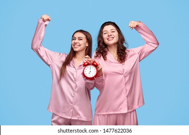 Cheerful Teen Girls In Pajamas Holding Alarm Clock And Flexing Biceps While Being Awake Early In Morning Against Blue Background