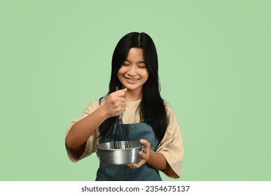 Cheerful teenage Asian girl wearing apron holding whisk and bowl isolated on green background - Powered by Shutterstock