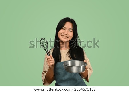 Cheerful teenage Asian girl wearing apron holding whisk and bowl isolated on green background