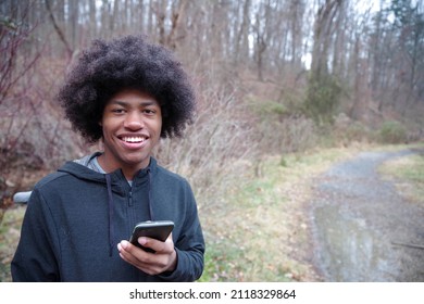 Cheerful Teenager Holds His Phone By A Wooded Hiking Trail With A Wooded Background. Shot In Natural Light With One Person, And Copy Space.