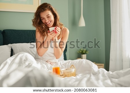 Cheerful tender female in white sleepwear sitting with cup of coffee on bed and having breakfast while enjoying morning in bedroom at home