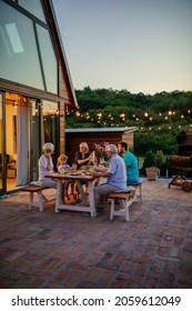 Fröhliche Familie der drei Generationen, die zusammen um einen Tisch im Garten zu Abend essen. Der Garten ist mit Lampen dekoriert