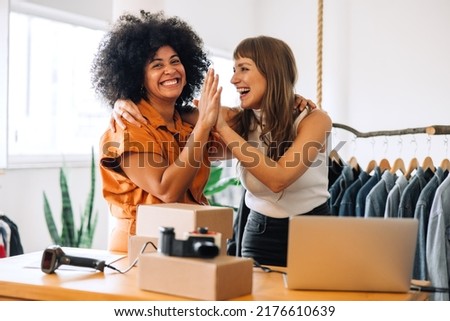 Cheerful thrift store owners high-fiving each other in their shop. Happy young businesswomen celebrating their success as a team. Two female entrepreneurs running an e-commerce small business.