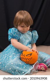 A Cheerful Toddler Girl In A Beautiful Blue Dress Takes Out Candy From A Pumpkin. Jack-O-Lantern