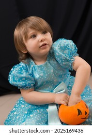 A Cheerful Toddler Girl In A Beautiful Blue Dress Takes Out Candy From A Bucket Of Pumpkins. Jack-O-Lantern