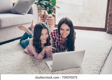 Cheerful toothy smiling happy mother and her little curious daughter are lying on a floor at home and usinf a laptop for watching cartoons, a girl is indicating on the monitor at the weekend - Powered by Shutterstock