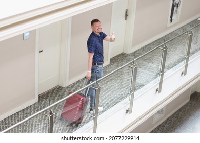 Cheerful tourist man with suitcase search for his room in hotel show thumbs up gesture