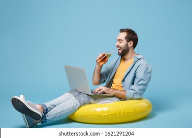 Cheerful Traveler Tourist Man In Yellow Clothes Isolated On Blue Background. Passenger Traveling Abroad On Weekend. Air Flight Journey Concept. Sit In Inflatable Ring Work On Laptop, Hold Beer Bottle