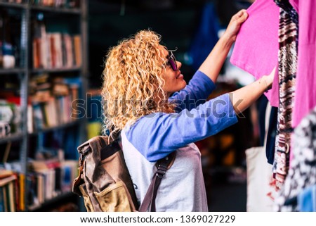 Cheerful traveler young blonde curly woman looking and choosing clothes at the used market during alternative vacation - shopping lifestyle concept for nice people