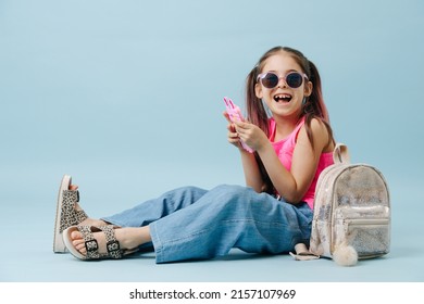 Cheerful Tween Girl In Pink Shirt And Round Sunglasses Using Her Phone. Sitting On The Floor Over Blue Background.