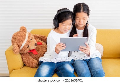 Cheerful two little kid sisters sit at yellow couch enjoy watching online movies or playing games on wireless tablet computer. Cute child with headset and her sister looking at ipad screen together.